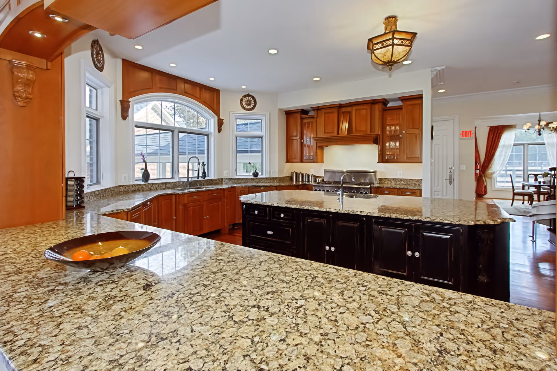 Bright, spacious kitchen featuring granite countertops, a large island with sink, wooden cabinetry, and arched windows.