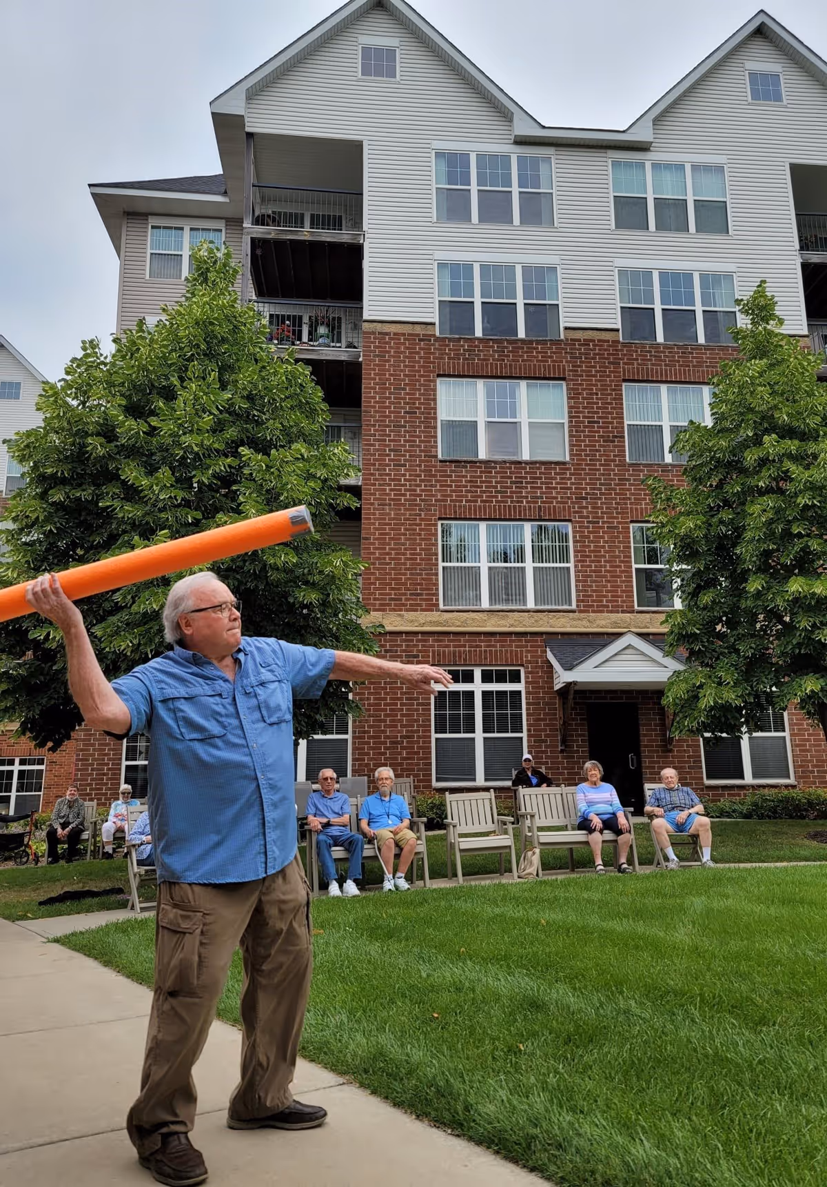An elderly man in a blue shirt throws an orange foam noodle on the lawn in front of a multi-story brick-and-siding senior living building with other residents seated on benches.