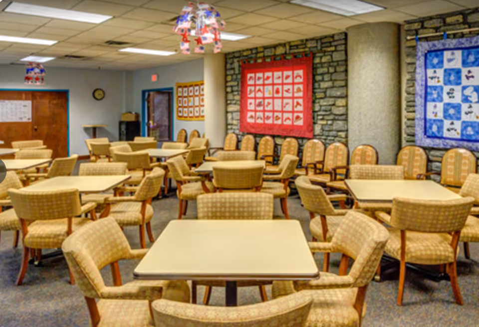 A spacious dining room in The Lodge Retirement Community with multiple square tables surrounded by cushioned chairs. The walls feature colorful quilts and stone accents, and the ceiling has fluorescent lighting with decorative hanging lamps.