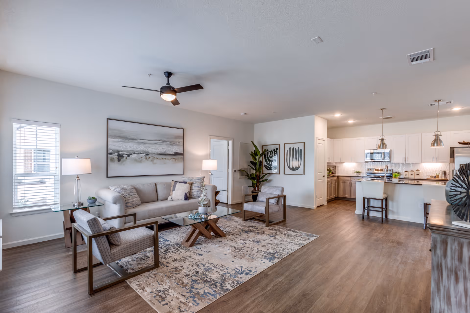 A modern open-concept living room and kitchen area with light gray walls and wood flooring. The living room features a beige sofa with multiple cushions, two armchairs with wooden frames, a glass coffee table on a patterned rug, and two table lamps on side tables. The kitchen has white upper cabinets, wood lower cabinets, stainless steel appliances, and a breakfast bar with two chairs. There are two pendant lights hanging over the kitchen island, and decorative artwork and plants are visible on the walls and surfaces.