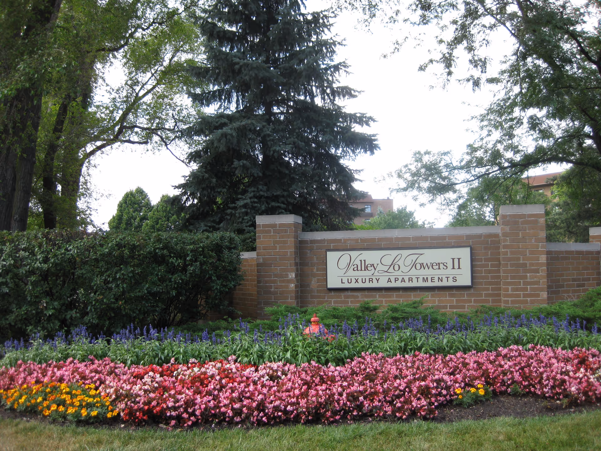 A brick sign for Valley Lo Towers II Luxury Apartments surrounded by colorful flower beds and greenery, with tall trees in the background.