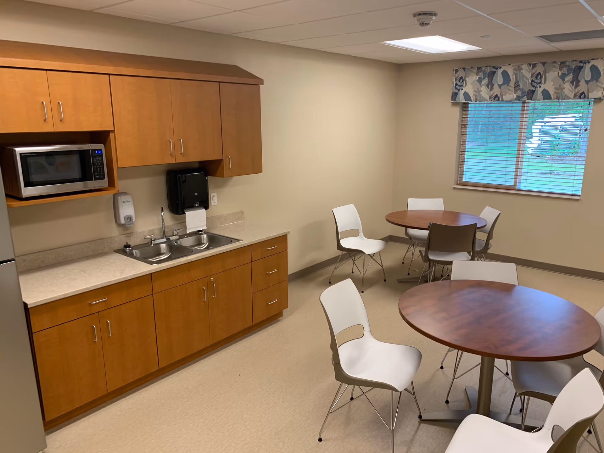A small kitchen and dining area with wooden cabinets, a microwave, a double sink, and a refrigerator on the left side. There are two round wooden tables with white chairs around them. A window with blinds and a floral valance is on the right wall, letting in natural light.