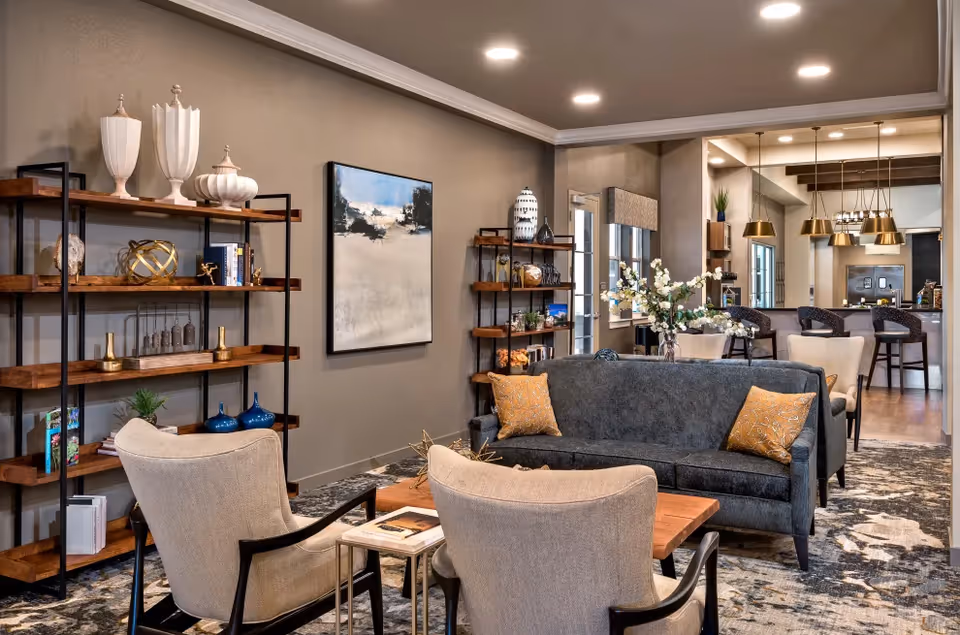 A modern and cozy living room area with a dark gray sofa adorned with two orange patterned pillows, two beige armchairs, and a wooden coffee table. Behind the sofa, there are two wooden shelves with decorative items and books. The room features a large mirror on the far wall reflecting a kitchen area with pendant lights and bar stools. The walls are painted in a neutral tone, and the floor is covered with a patterned rug.