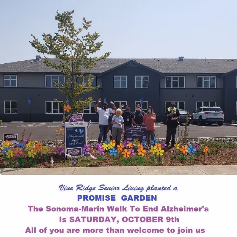 A group of seven people standing outside in front of a two-story building with a parking lot. They are behind a garden bed filled with colorful flower decorations and several signs. The people are wearing masks and some are waving. The sky is clear and the scene appears to be a community event.