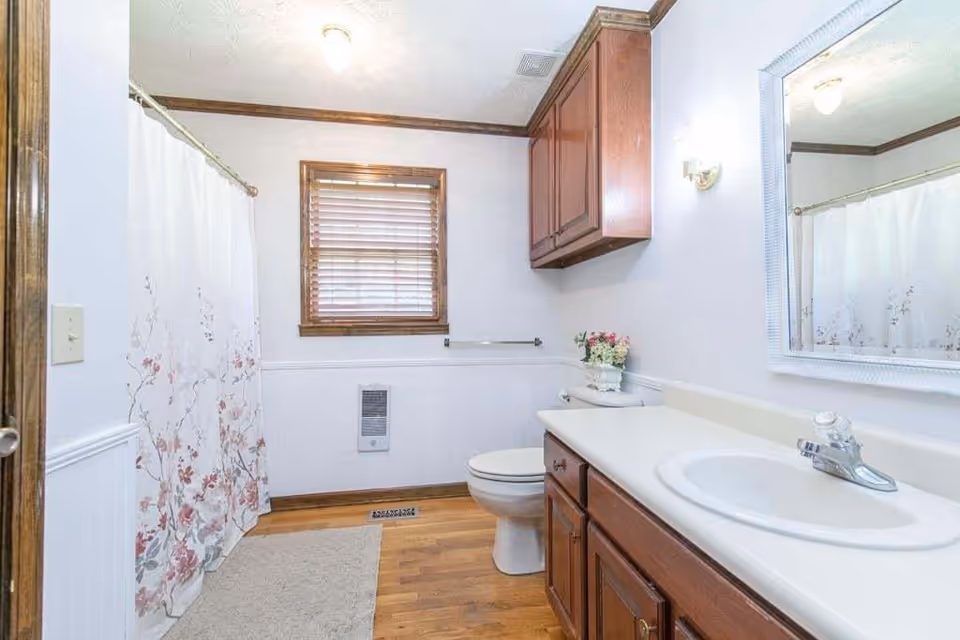 A clean bathroom featuring a white sink with a chrome faucet set in a beige countertop, wooden cabinets below and above the toilet, a white toilet, a window with wooden blinds, a floral shower curtain, a beige bath mat on a wooden floor, and a wall-mounted heater beneath the window.