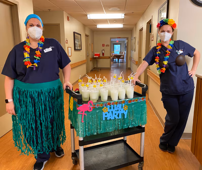 Two healthcare workers wearing navy scrubs, face masks, and colorful Hawaiian-themed accessories stand in a hallway. They are posing with a decorated cart filled with multiple cups of a light-colored drink with straws and garnishes. The cart is decorated with green fringe, a pink flamingo cutout, and a sign that reads 'Luau Party'.