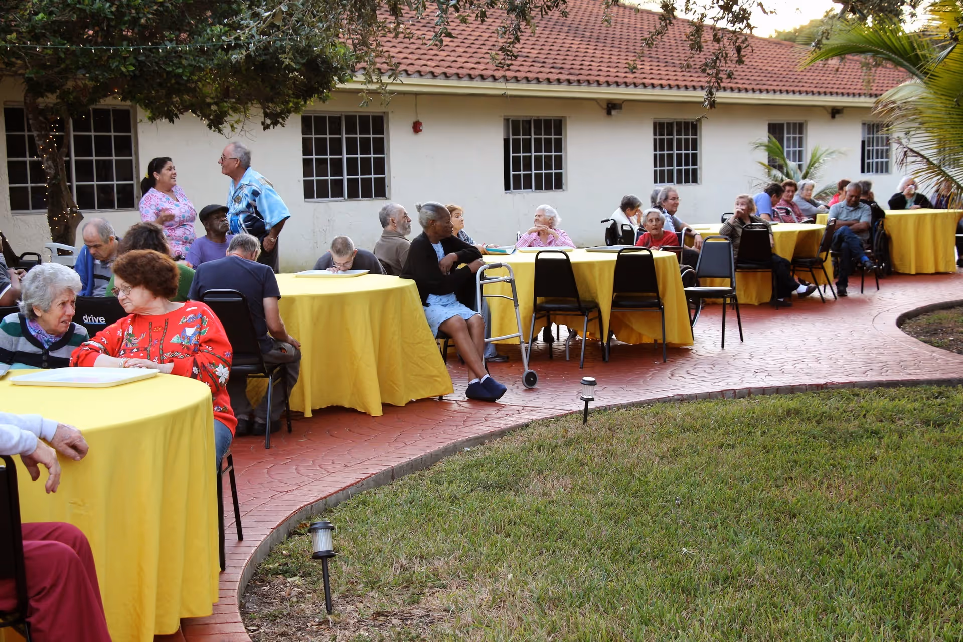 A group of elderly people sitting and socializing at round tables covered with yellow tablecloths outside a building with white walls and red roof tiles. The area is paved with red bricks and surrounded by grass and trees.