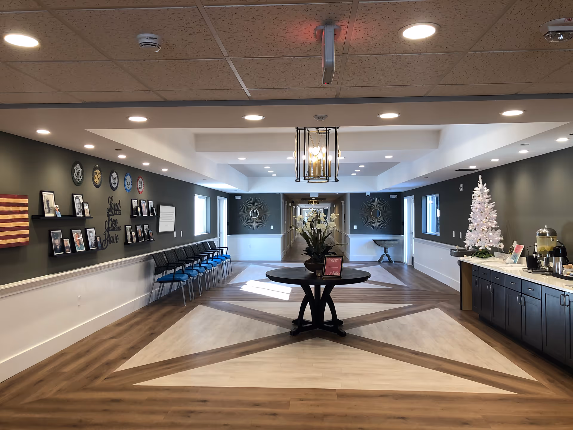 Interior view of a senior living facility hallway with a central round table decorated with flowers. On the left wall, there are framed photos and military insignias with the words 'Land of the Free Brave'. Chairs are lined up along the wall. On the right side, there is a counter with a white Christmas tree and refreshments. The floor has a geometric wood pattern, and the ceiling has recessed lighting and a hanging light fixture.