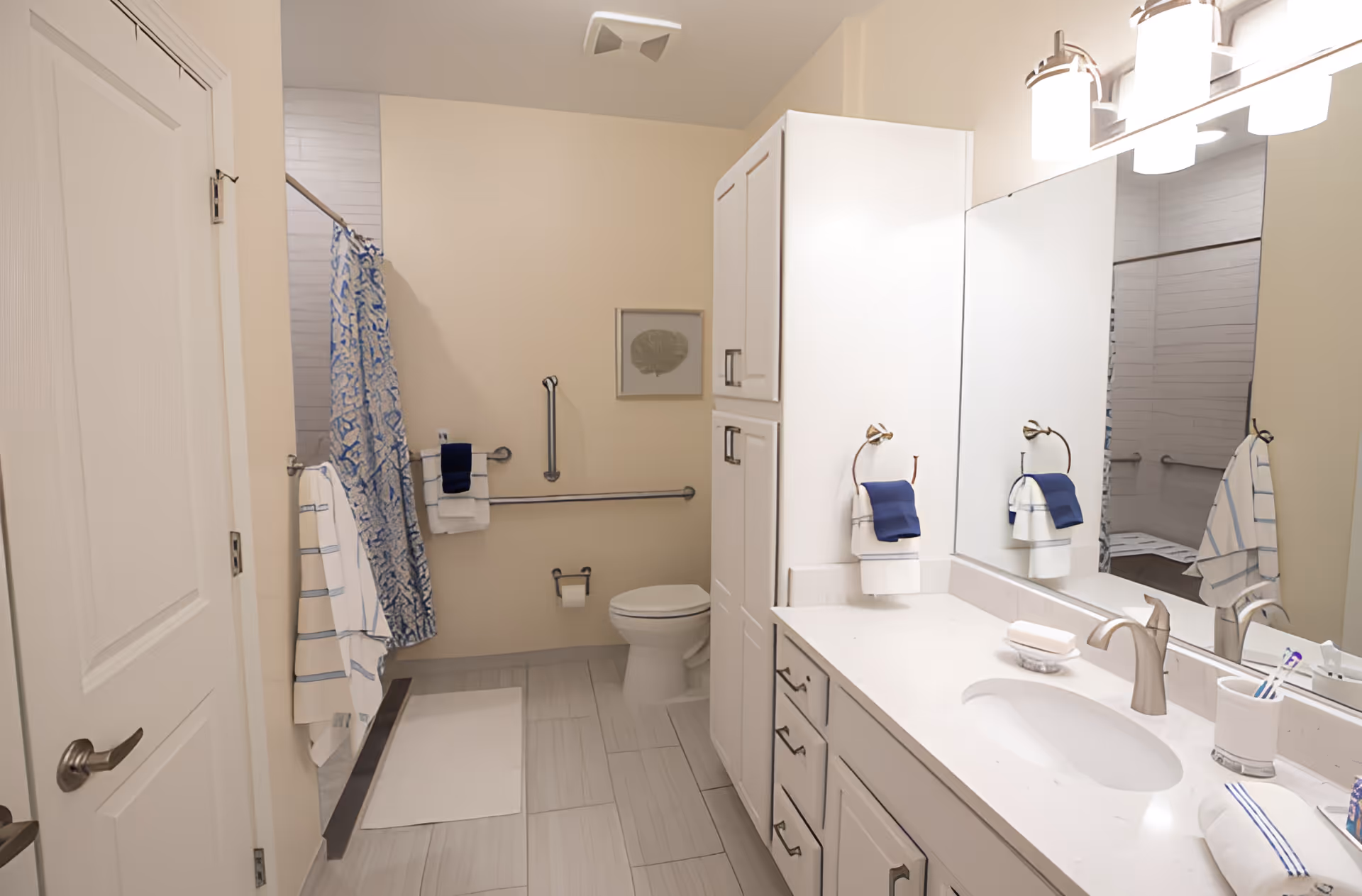 A clean and modern bathroom featuring a white vanity with a sink, a large mirror, and silver fixtures. There is a toilet next to a wall with grab bars, a shower area with a blue and white patterned curtain, and several towels hanging on hooks and bars. The floor is tiled in light gray, and the walls are painted a soft beige color.