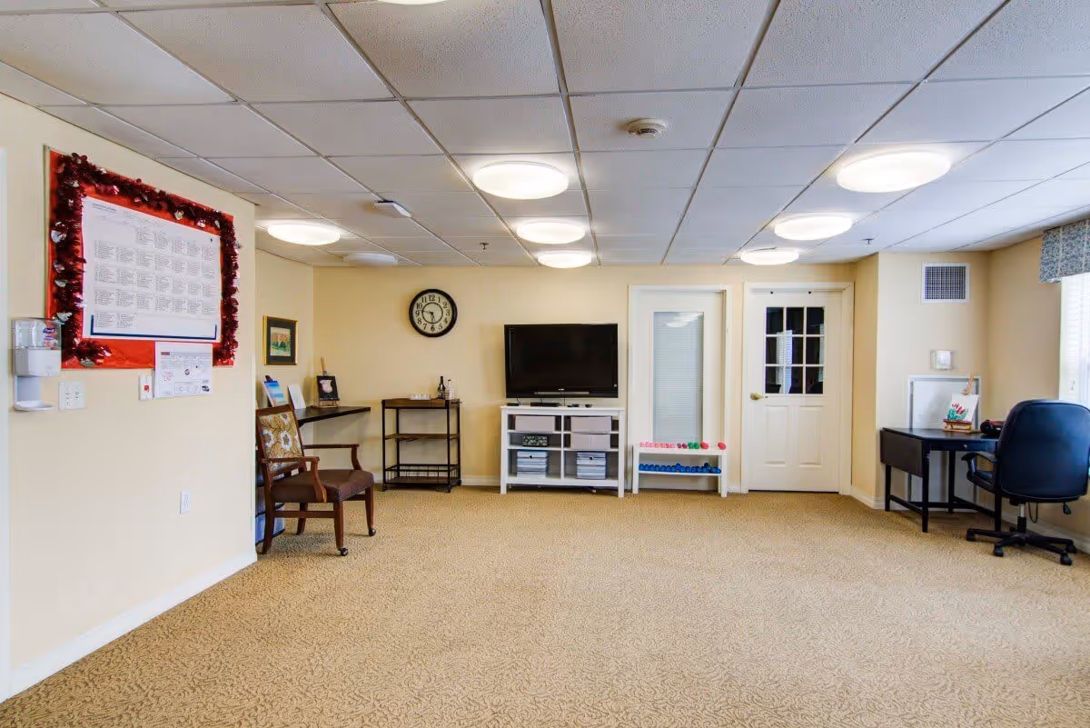 A spacious room with beige walls and carpet, featuring a wall-mounted clock, a flat-screen TV on a white stand with storage bins, a wooden chair, a small desk with a black office chair, and two doors. The ceiling has multiple round light fixtures, and there is a bulletin board decorated with red garland on the left wall.