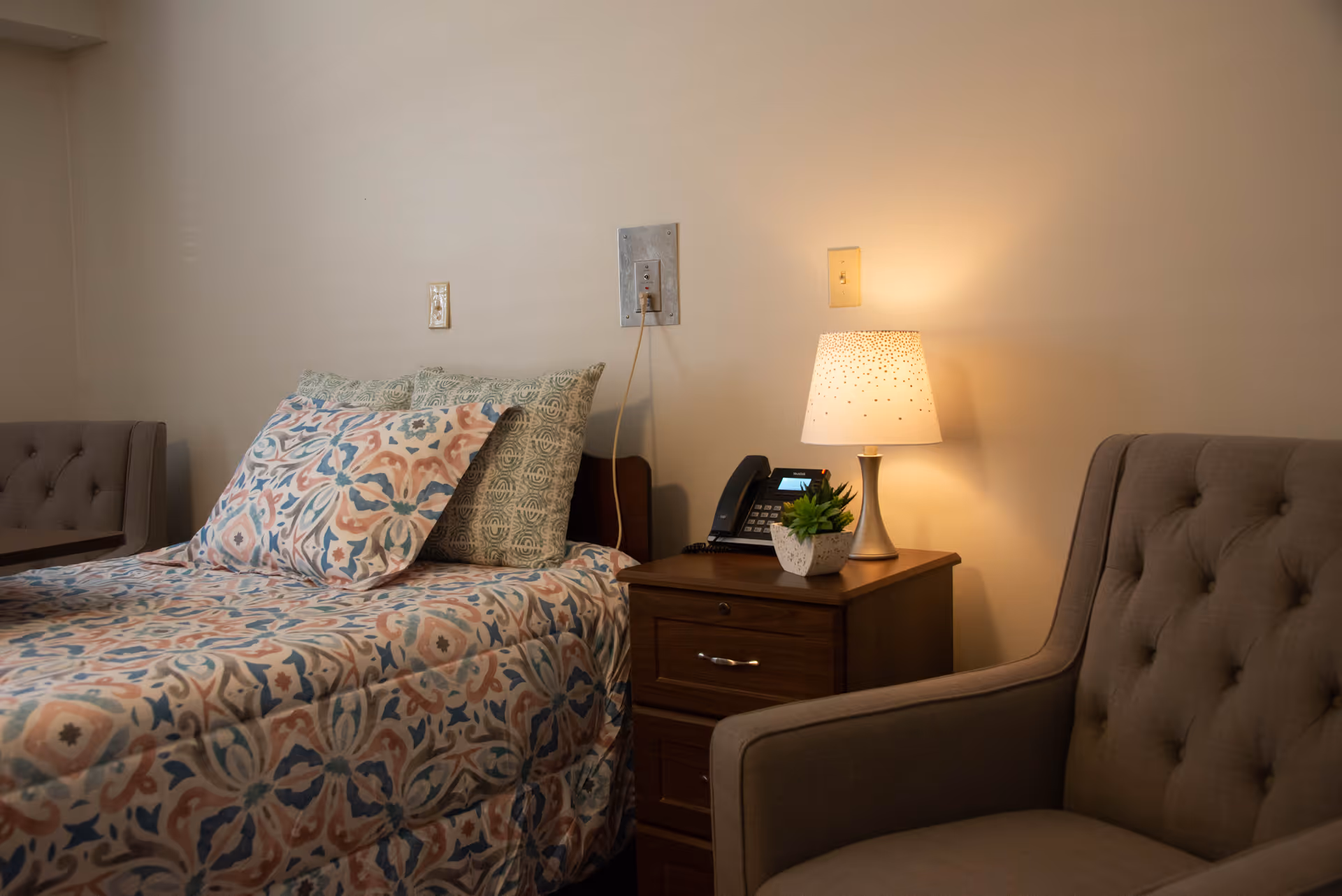 A neatly made single bed with patterned bedding, a wooden nightstand holding a lamp, phone and small plant, and an upholstered chair in a bedroom.