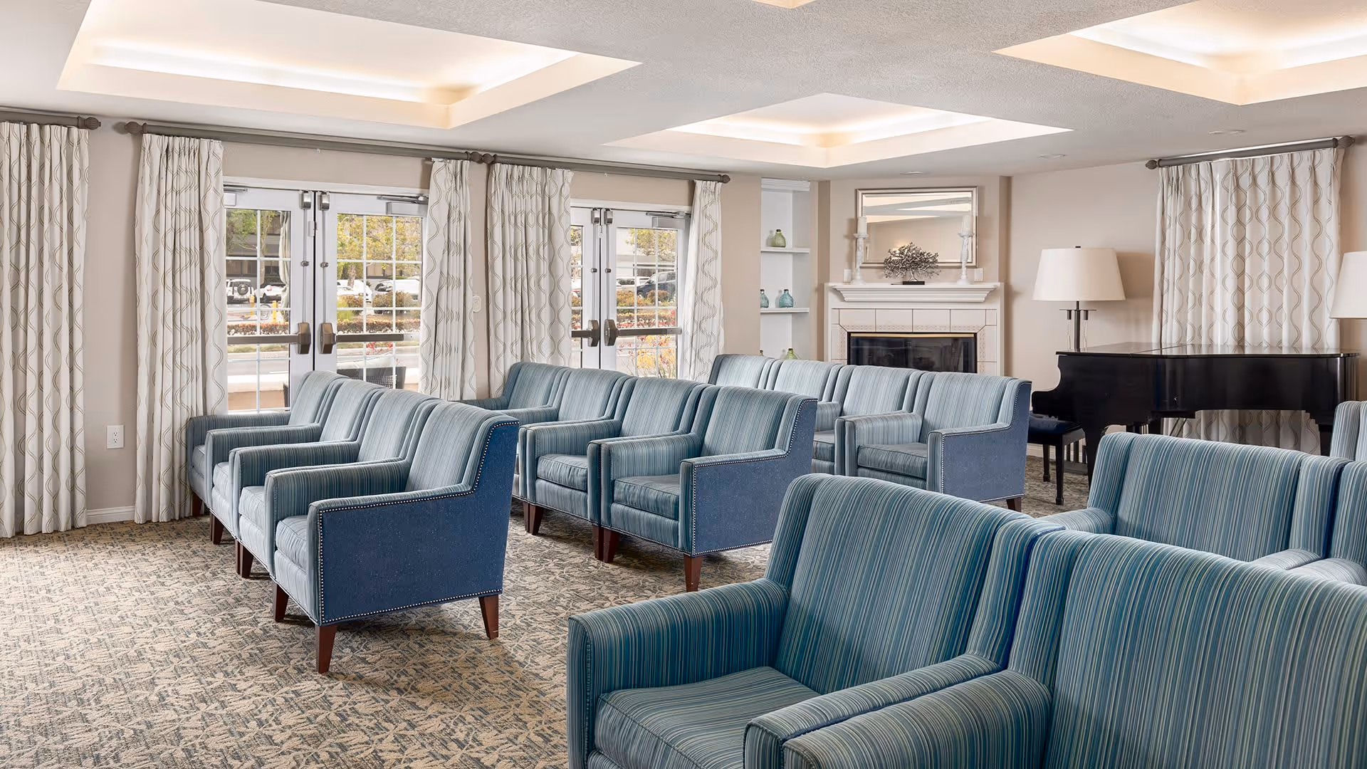 A bright and spacious living room with multiple blue upholstered armchairs arranged in rows facing a fireplace. The room features large windows with patterned curtains, a grand piano in the corner, and soft overhead lighting.
