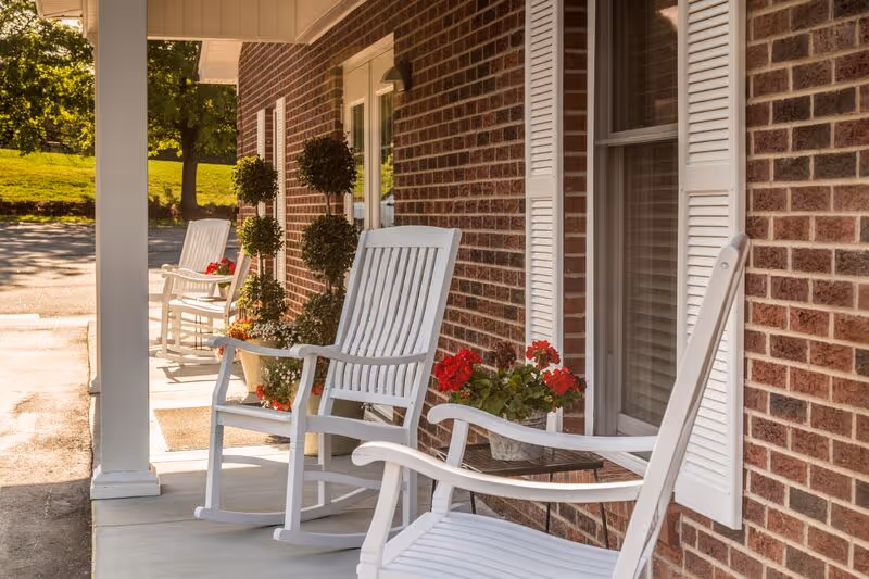 Front porch of a brick building with white rocking chairs and potted red flowers.