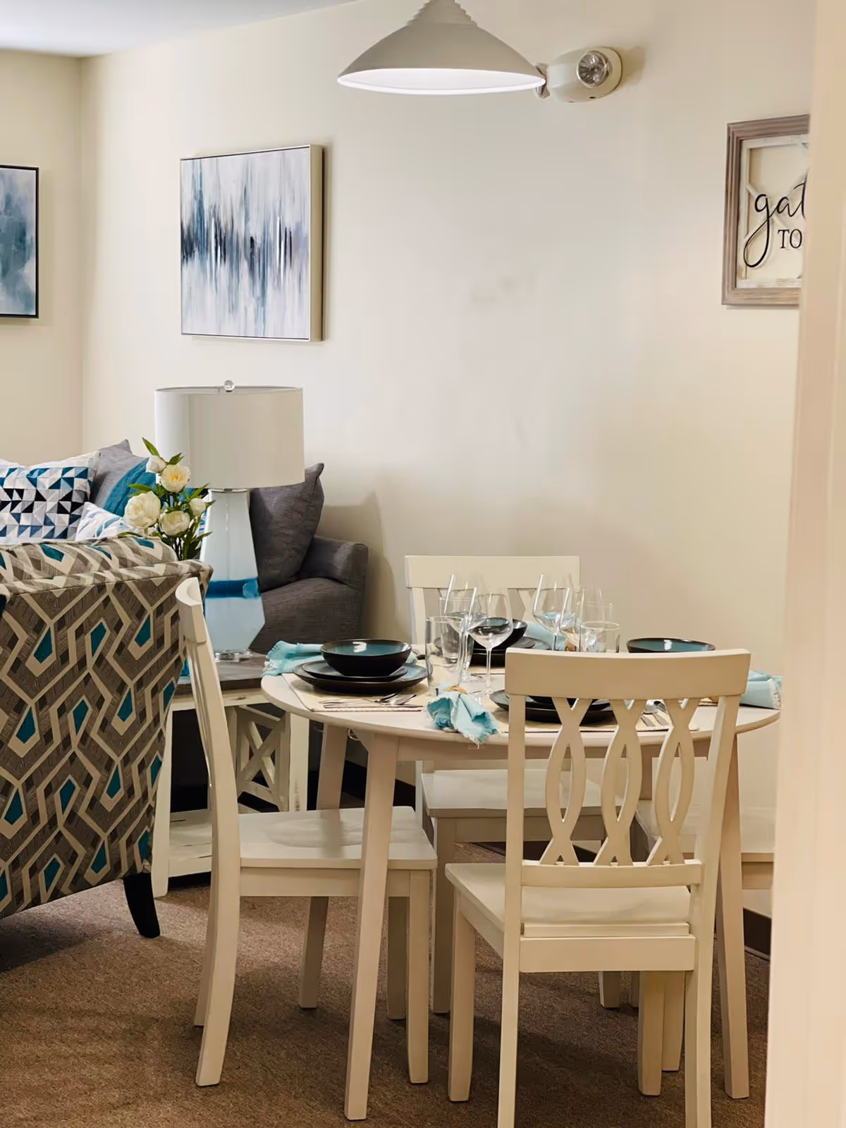 A cozy dining area with a round white table set for four with plates, bowls, glasses, and turquoise napkins. Surrounding the table are four white chairs with decorative backs. In the background, there is a gray couch with patterned cushions, a white table lamp, and abstract wall art on a light-colored wall.