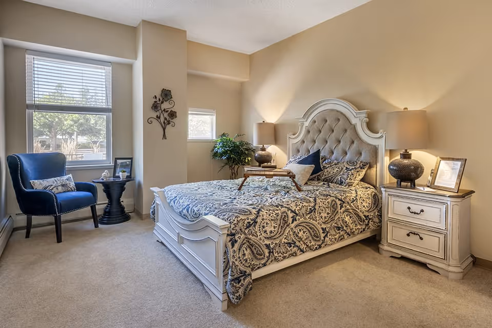 A cozy bedroom with a large bed featuring a tufted headboard and patterned bedding. There are two bedside tables with lamps, a small plant in the corner, a blue armchair with a decorative pillow, and a round black side table near two windows with blinds.
