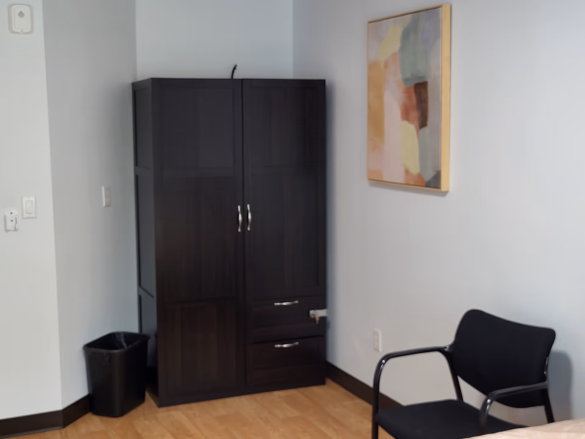 A corner of a room with a dark wooden cabinet, a black trash bin, a black chair with metal armrests, and an abstract painting on a light gray wall. The floor is wooden.