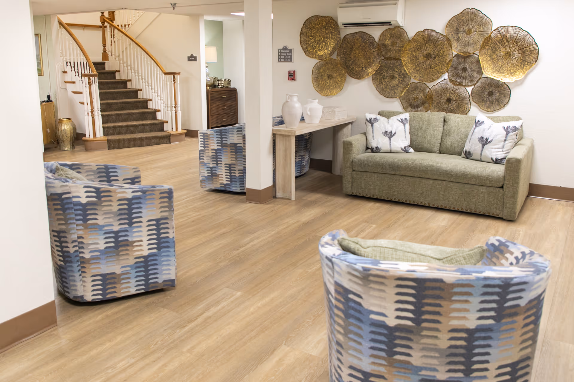 A cozy senior living facility common area with a green sofa adorned with floral pillows, three patterned armchairs, a wooden floor, and a decorative wall art installation made of circular metallic pieces. A staircase with wooden handrails is visible in the background.