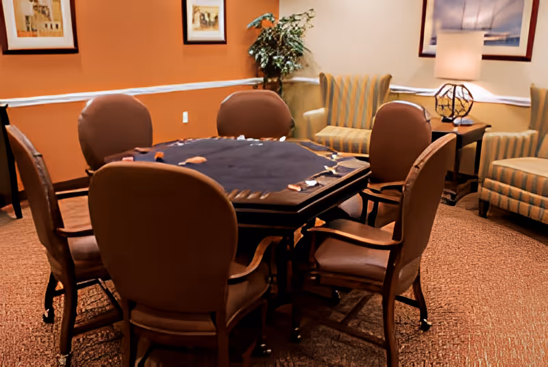 Cozy lounge area with a felt-covered square game table surrounded by six upholstered chairs and additional armchairs and a lamp.