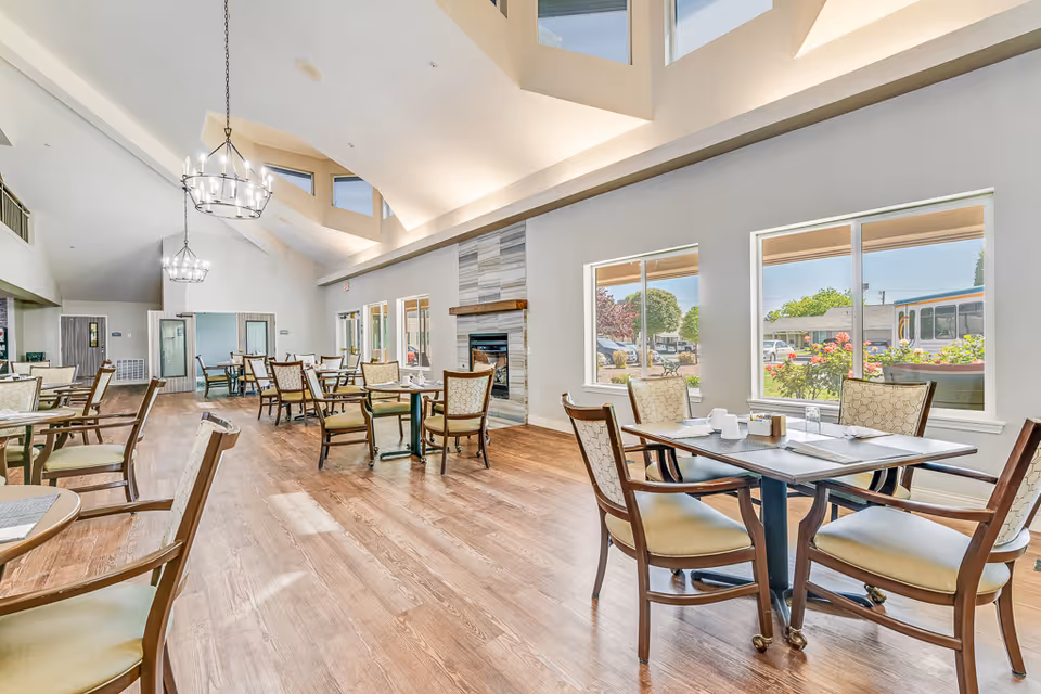 Bright and spacious dining room with wooden floors, multiple tables and chairs arranged neatly, large windows letting in natural light, and a modern fireplace on one wall. The ceiling is vaulted with hanging chandeliers, and the room has a welcoming atmosphere.
