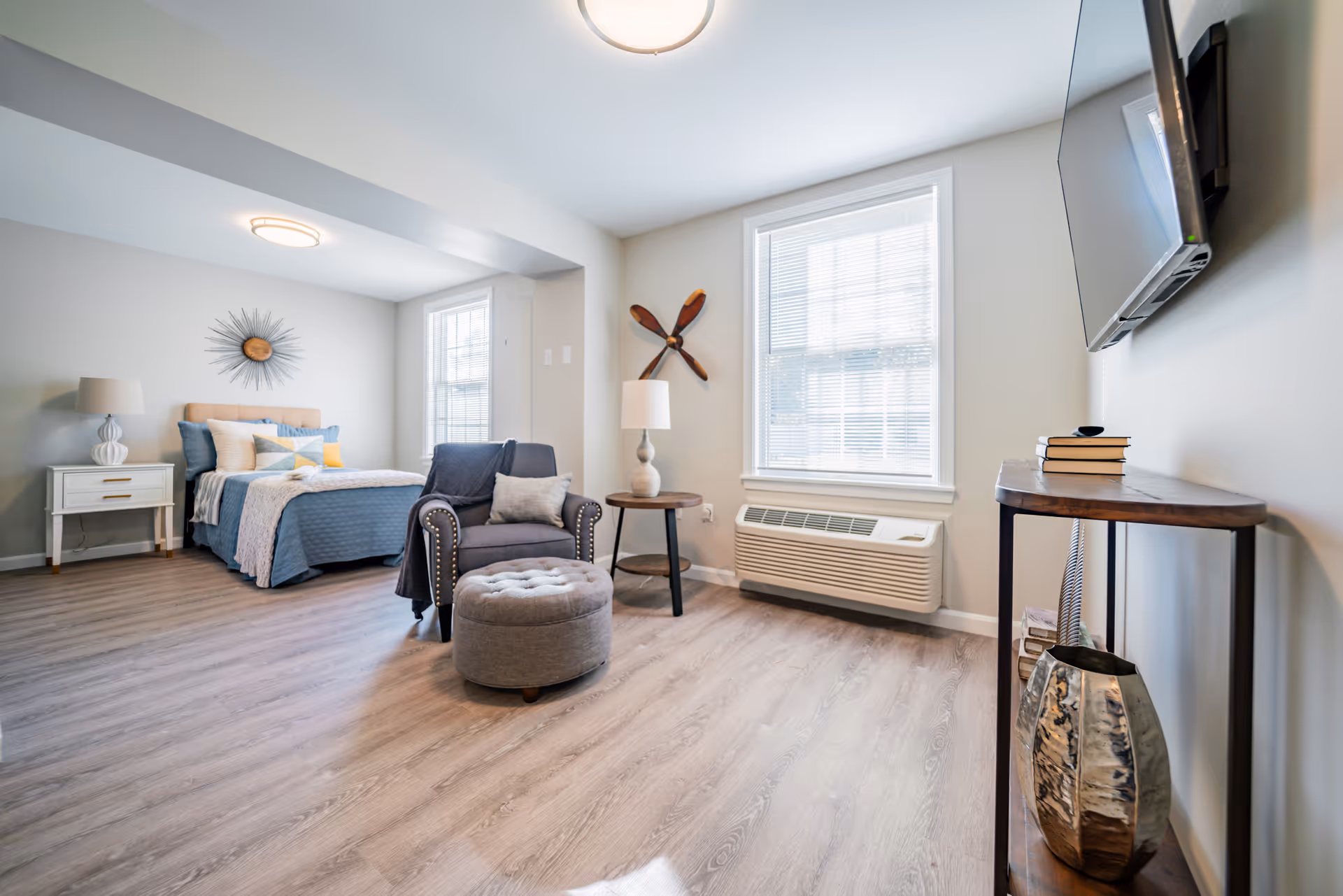 A bright and spacious bedroom with light wood flooring, a bed with blue and beige bedding, a gray armchair with a matching ottoman, a side table with a lamp, a wall-mounted TV, and decorative items including a sunburst wall clock and wooden propeller on the walls.
