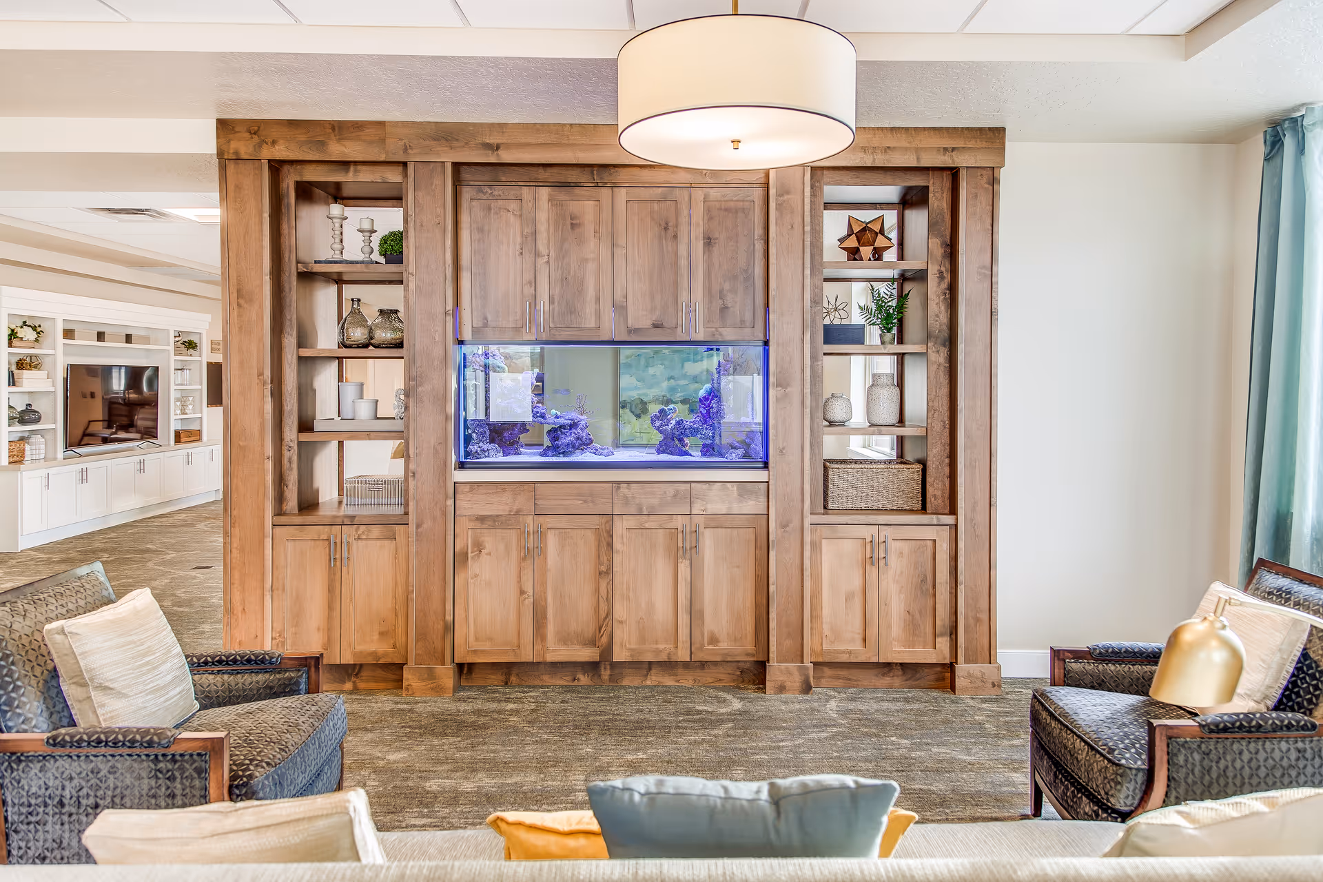 A cozy living room area featuring a large wooden built-in cabinet with an aquarium in the center. The cabinet has shelves with decorative items and closed storage. Surrounding the cabinet are comfortable armchairs with cushions, a hanging ceiling light, and a glimpse of another room with a TV and more cabinetry in the background.