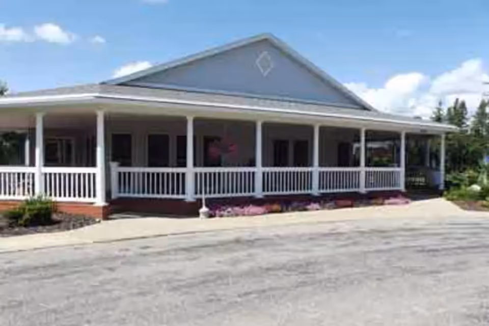 Single-story building with a wraparound porch featuring white railings and columns, surrounded by a paved driveway and some landscaping with purple flowers and greenery under a blue sky with scattered clouds.