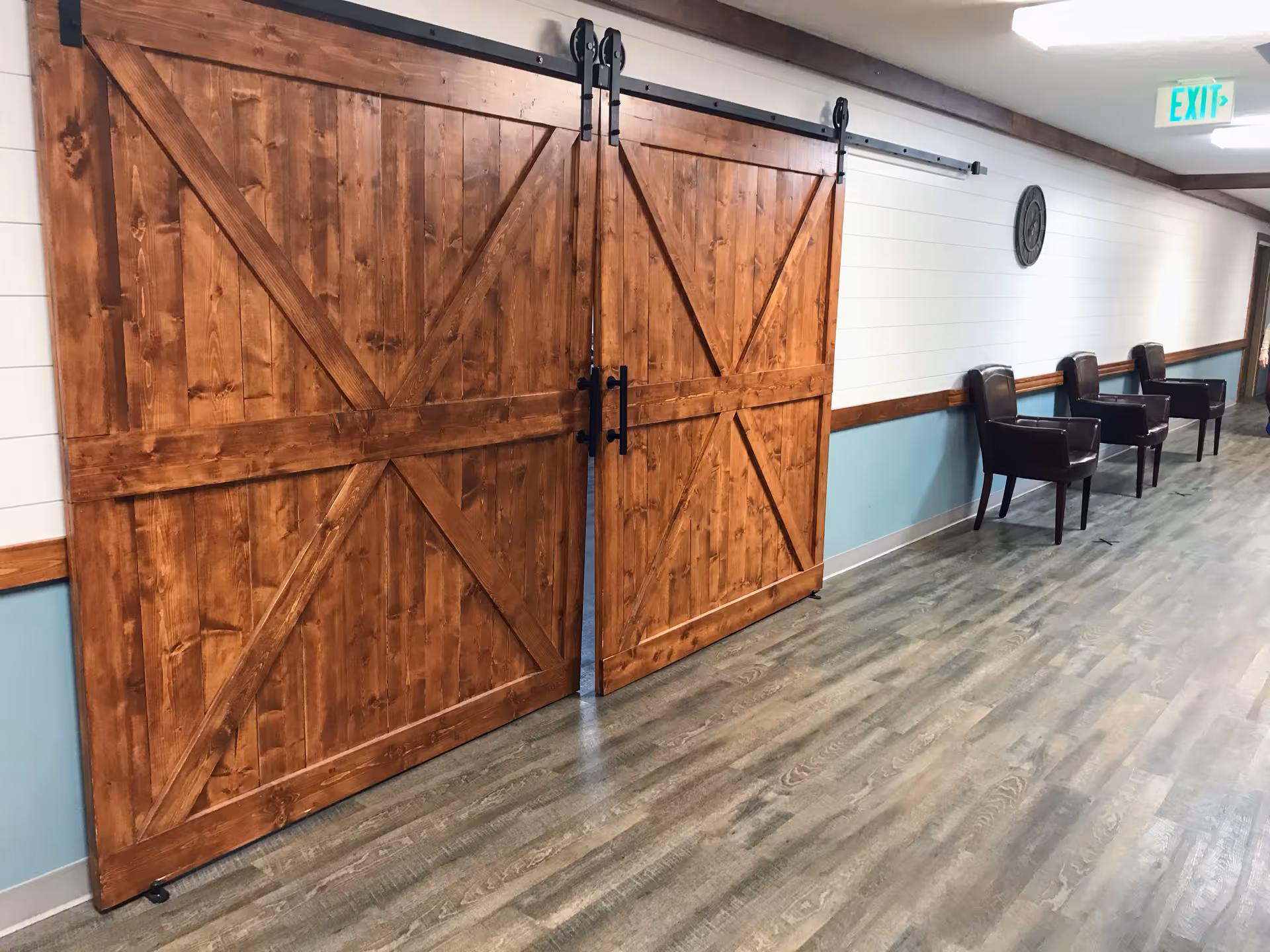 A hallway in a senior living facility with wooden sliding barn doors on the left and four dark brown chairs lined up against the right wall. The walls are painted white on the upper half and light blue on the lower half with a wooden trim separating the colors. There is an exit sign visible at the end of the hallway.