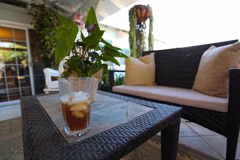 Outdoor patio area with a dark wicker sofa with beige cushions and a glass-top wicker table holding a glass of iced tea and a potted plant. The background shows a covered patio with hanging plants and a sliding glass door.