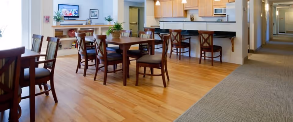 A bright and clean common area in a senior living facility featuring wooden tables and chairs on a hardwood floor. There is a kitchen area with a counter and bar stools, and a hallway with carpet leading to other rooms. The room has light-colored walls and some plants and decorations on shelves.