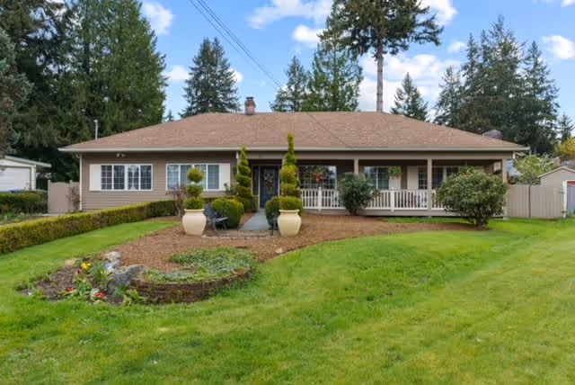 Single-story house with a brown roof and beige siding, surrounded by a well-maintained green lawn and landscaped garden beds. The front porch has white railings and is decorated with potted plants and shrubs. Tall trees are visible in the background under a partly cloudy sky.