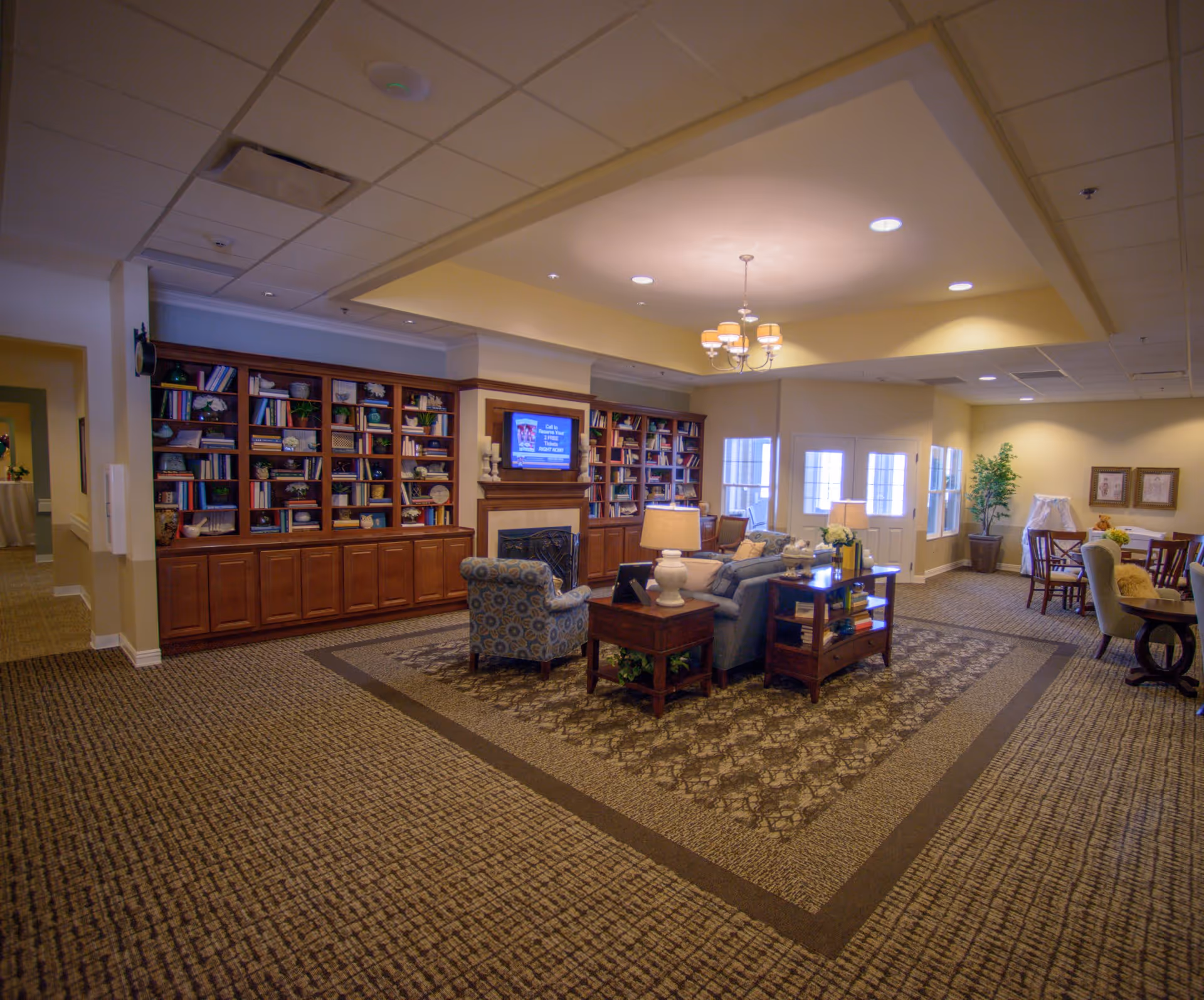 A spacious and well-lit living room area in a senior living facility featuring a patterned carpet, a built-in wooden bookshelf filled with books and decorative items, a fireplace with a TV mounted above it, comfortable seating including an armchair and a sofa, side tables with lamps, and a dining area with tables and chairs in the background.