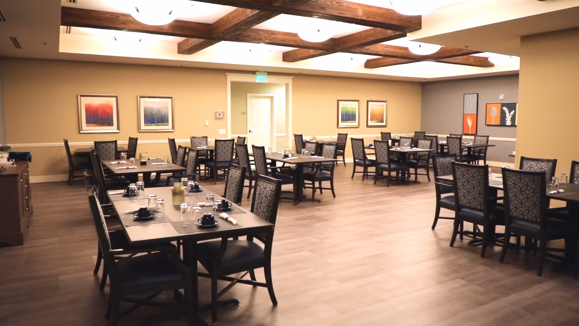 A spacious dining room with multiple dark wood tables and patterned chairs arranged neatly. Each table is set with black cups, glasses, and napkins. The room features wooden ceiling beams, light-colored walls with framed artwork, and a wooden floor.
