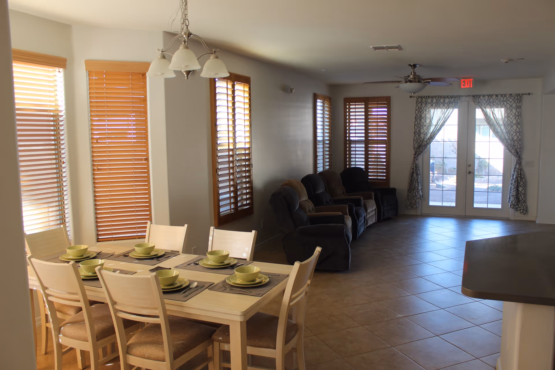 A bright interior room with a dining table set for six with green bowls and plates, beige chairs, and wooden blinds on the windows. In the background, there are several recliner chairs lined up against the wall and a set of glass double doors with patterned curtains leading outside.