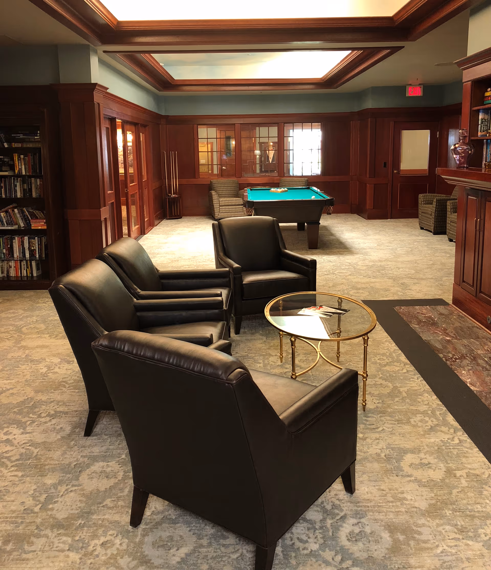 A cozy common area with four black leather armchairs arranged around a small round glass coffee table with a gold frame. In the background, there is a pool table with two patterned armchairs on either side. The room features wood-paneled walls, built-in bookshelves filled with books, and a carpeted floor with a subtle pattern. The ceiling has recessed lighting and a decorative wooden trim.