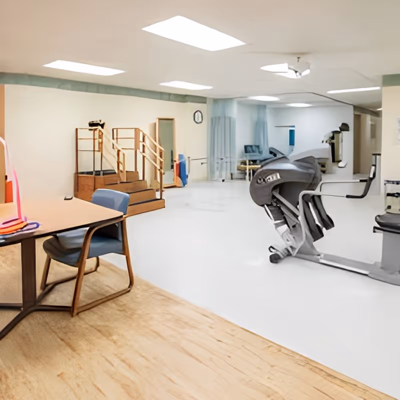 Bright rehabilitation therapy room with exercise equipment, parallel bars, therapy stairs, and a table with chairs.