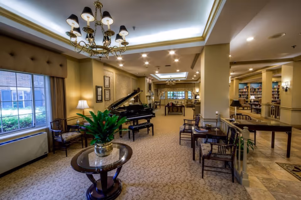 A spacious and elegant living room area in a senior living facility with beige walls and carpet. The room features a grand piano, multiple seating areas with chairs and tables, a chandelier, large windows with curtains, and a bookshelf in the background. There is a round table with a potted plant in the foreground.