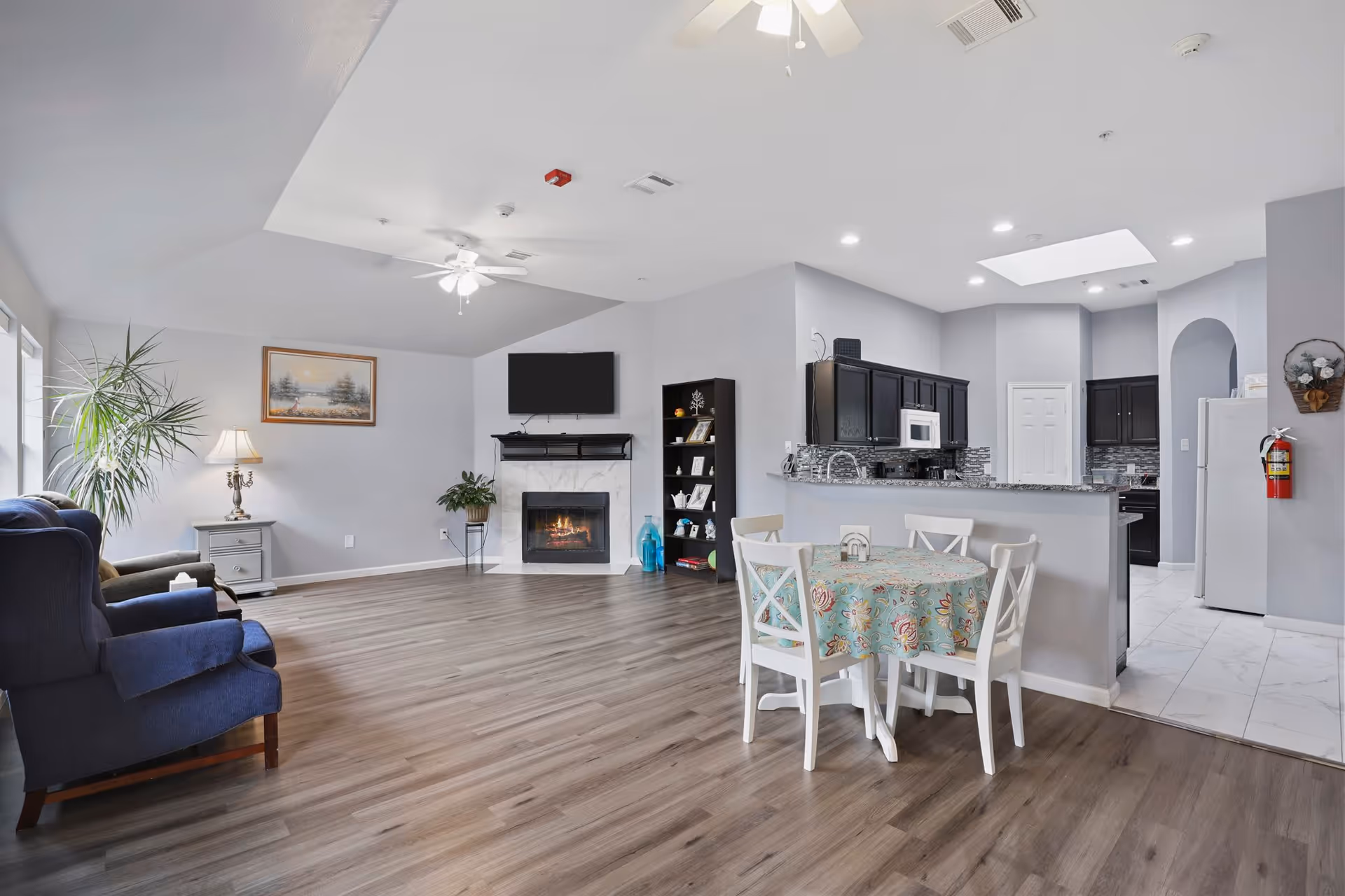 A spacious senior living common area featuring a seating area with two blue armchairs, a side table with a lamp, a large plant, a wall-mounted TV above a fireplace, and a bookshelf. Adjacent to the seating area is a small dining table with four white chairs and a floral tablecloth. The kitchen with dark cabinets, a microwave, and a refrigerator is visible in the background. The room has light gray walls, wood flooring, and ceiling fans with lights.