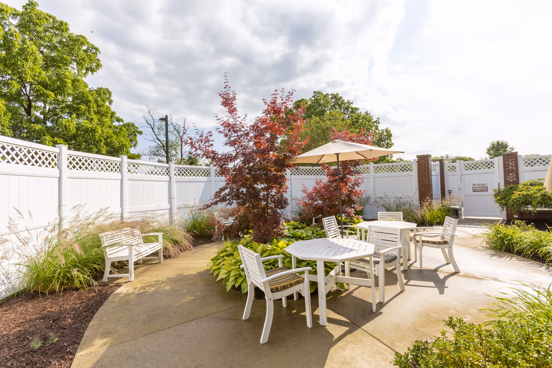 Outdoor patio area with white tables and chairs, some with cushions, surrounded by green plants and small trees with red leaves. A white fence encloses the space, and there is an umbrella providing shade over one of the tables. The sky is partly cloudy.