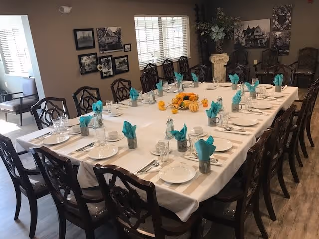 A large rectangular dining table set for a meal with white tablecloth, white plates, silverware, glasses, and turquoise folded napkins in metal holders. The table has a centerpiece with small pumpkins and autumn decorations. The room has wooden chairs with intricate backs, framed black and white photos on the walls, and a window with blinds letting in natural light.