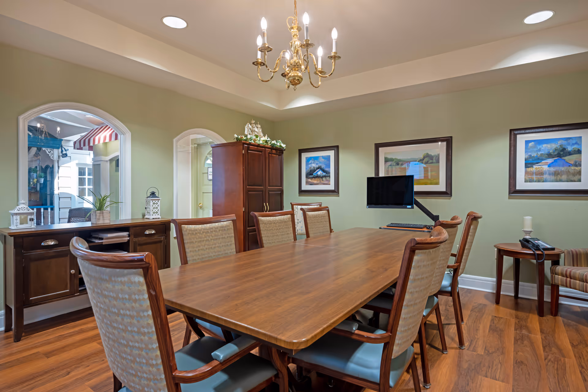 A well-lit conference or meeting room with a long wooden table surrounded by eight upholstered chairs. The room features light green walls adorned with three framed landscape paintings. A wooden cabinet and sideboard with decorative items are against the walls. A computer monitor and keyboard are placed on the table. The ceiling has a brass chandelier and recessed lighting. Two arched windows look into an adjacent room with a striped awning and plants.