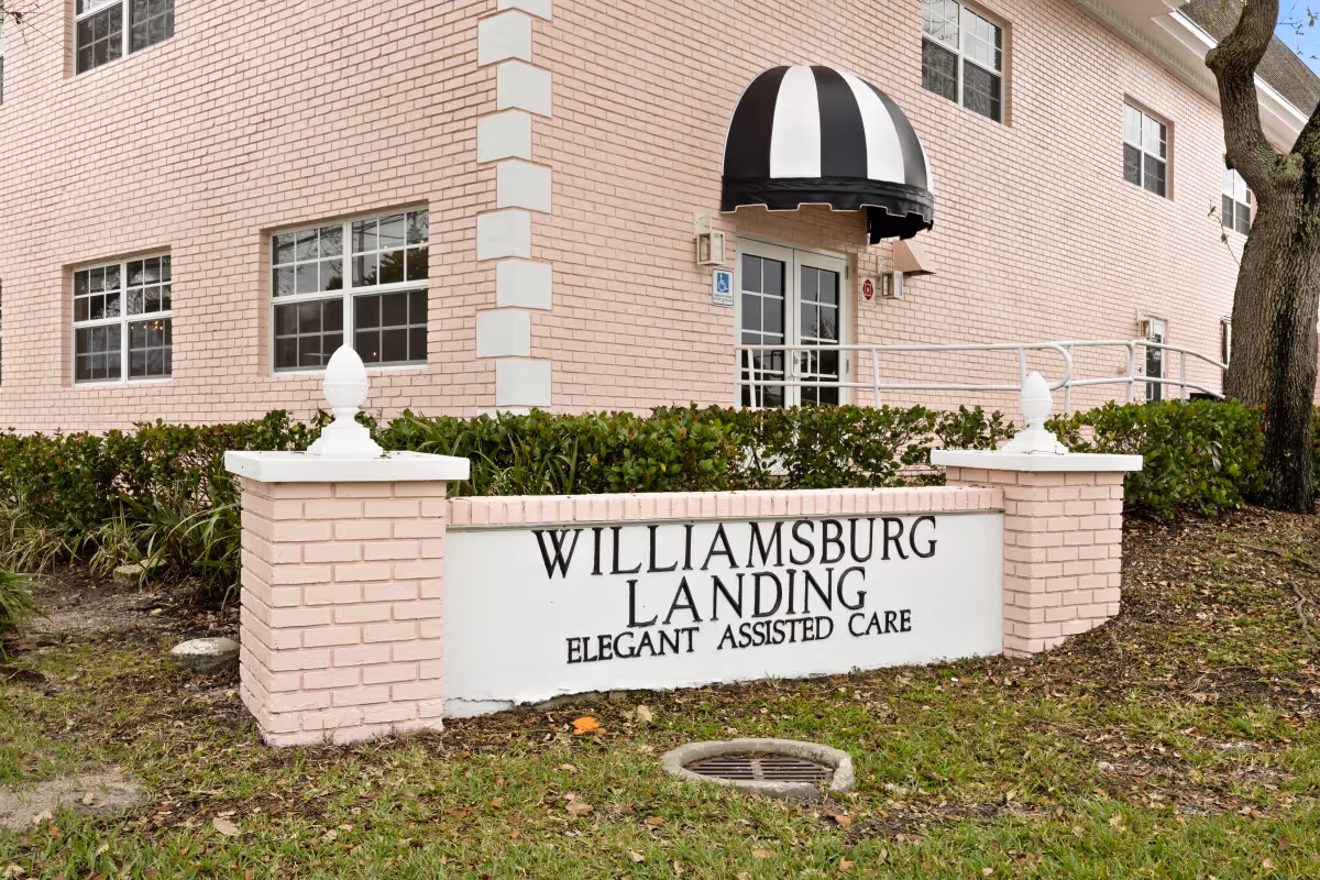 Exterior view of Williamsburg Landing Assisted Living facility showing a pink brick building with multiple windows and a black and white striped awning above the entrance door. In front of the building is a sign made of pink bricks and white panel that reads 'Williamsburg Landing Elegant Assisted Care'. There are green bushes along the building and a tree on the right side.