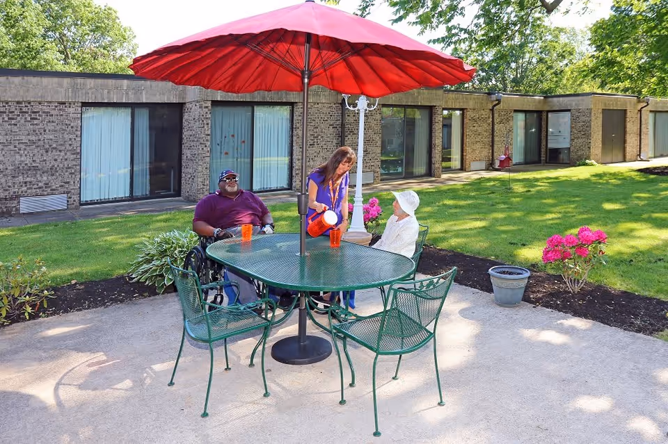 Two elderly individuals sitting at a green metal outdoor table with a large red umbrella. One person is in a wheelchair wearing a purple shirt and cap, and the other is seated wearing a white hat and light-colored clothing. A woman in a blue outfit is pouring a drink from an orange pitcher into a cup on the table. The setting is a sunny patio area with green grass, flowers, and a single-story brick building with large windows in the background.