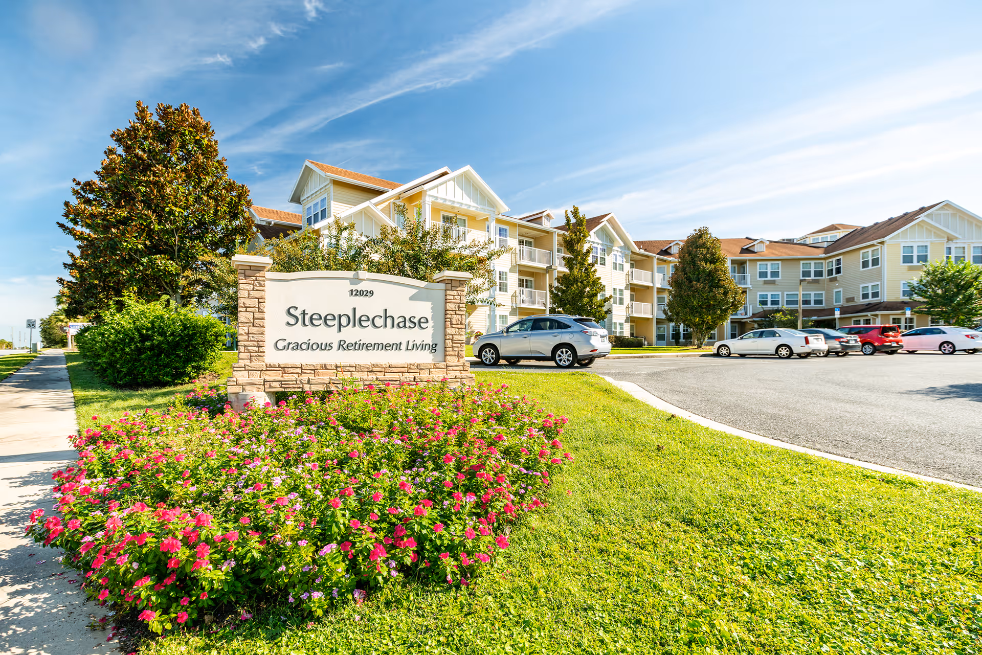 Exterior view of Steeplechase Gracious Retirement Living facility with a large sign in front surrounded by colorful flowers and green grass, a parking lot with several cars, and a multi-story residential building under a blue sky with light clouds.