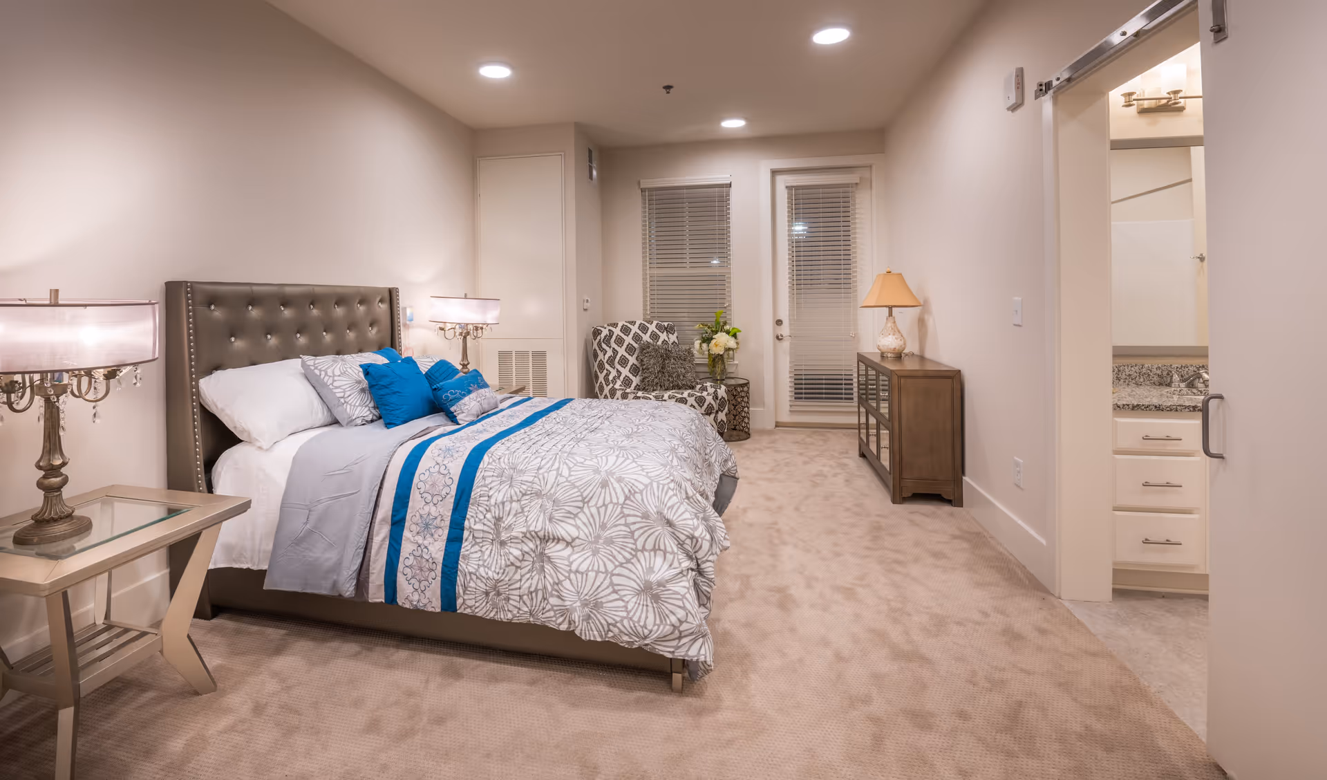 A neatly arranged bedroom with a large bed featuring a tufted headboard, white and blue bedding, and multiple pillows. There are two bedside tables with lamps, a patterned armchair near the window, a wooden dresser with a lamp, and a door leading to a bathroom with a granite countertop and white cabinets. The room has beige carpet and neutral-colored walls.