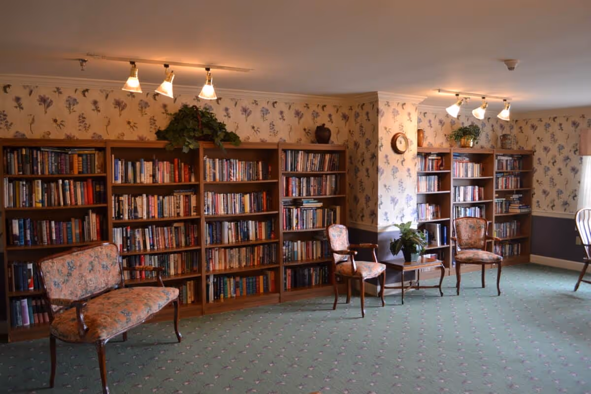 A cozy senior living facility library room with floral wallpaper and green carpet. The room features several wooden bookshelves filled with books, vintage-style upholstered chairs, a small wooden table with a potted plant, and ceiling track lighting.
