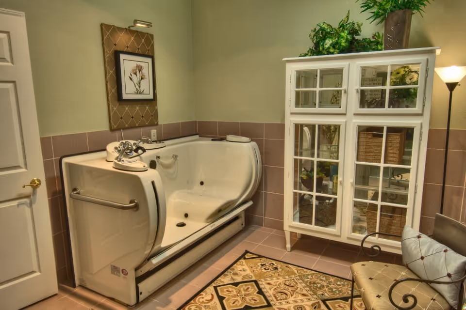 Tiled bathroom featuring a white walk-in bathtub, glass-front storage cabinet, and a cushioned chair on a patterned rug.