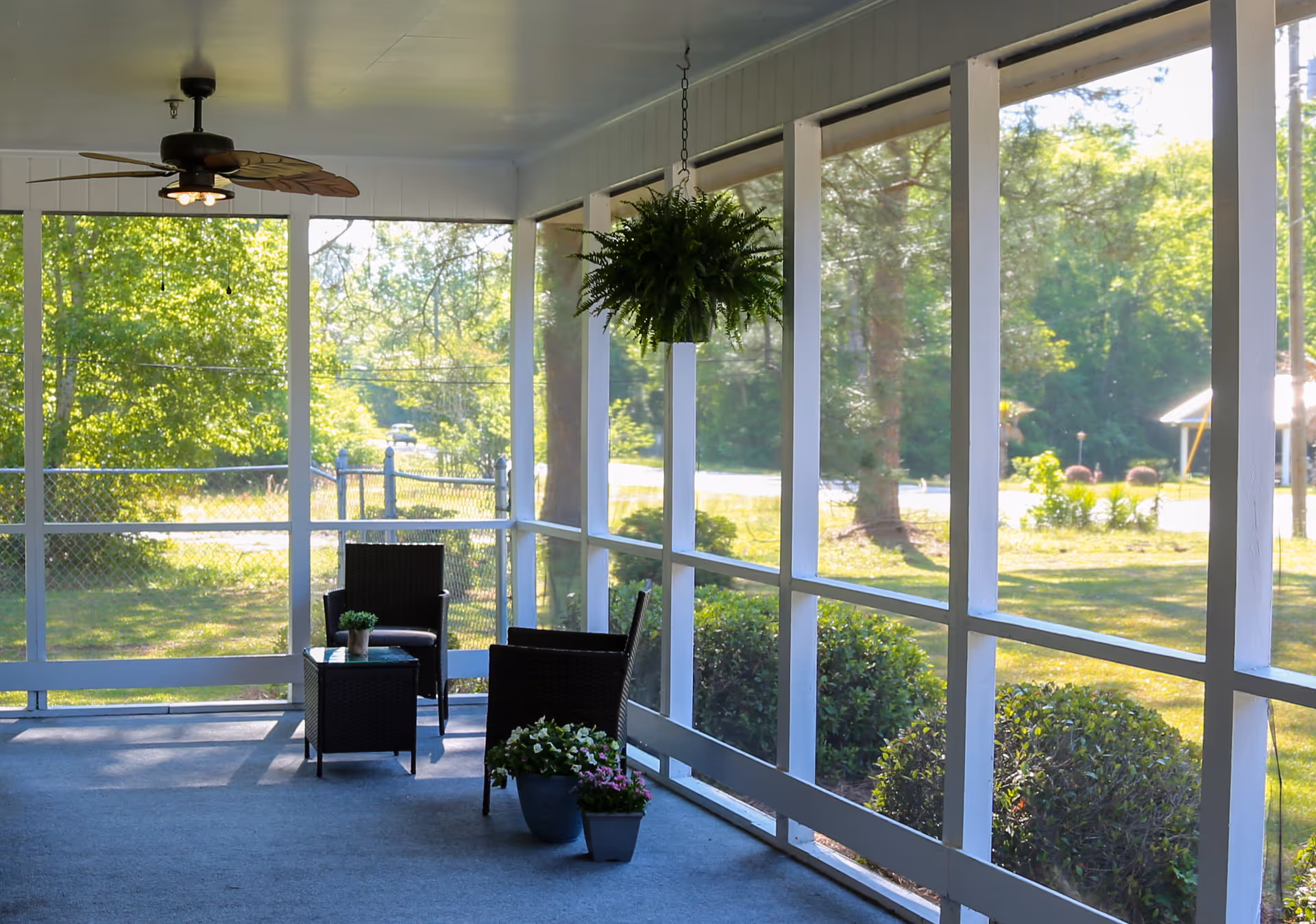 A screened-in porch with two black wicker chairs and a small table between them. There are potted plants on the floor and table, a hanging fern plant, and a ceiling fan with lights. Outside the porch, there is greenery including bushes, trees, and a lawn.