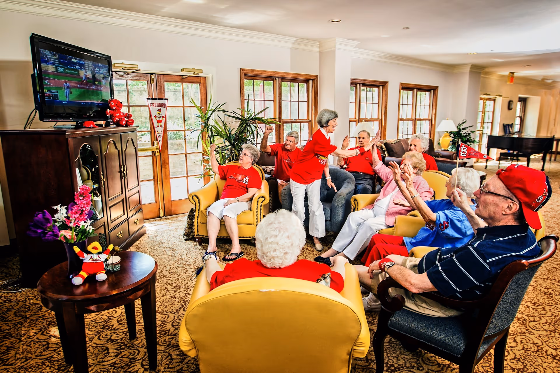 A group of elderly people sitting and standing in a bright living room area, watching a baseball game on a television. They are wearing red and blue clothing, some with baseball team logos, and appear to be cheering and interacting with each other. The room has large windows, yellow armchairs, a wooden cabinet with flowers and a stuffed toy on a side table, and a piano in the background.