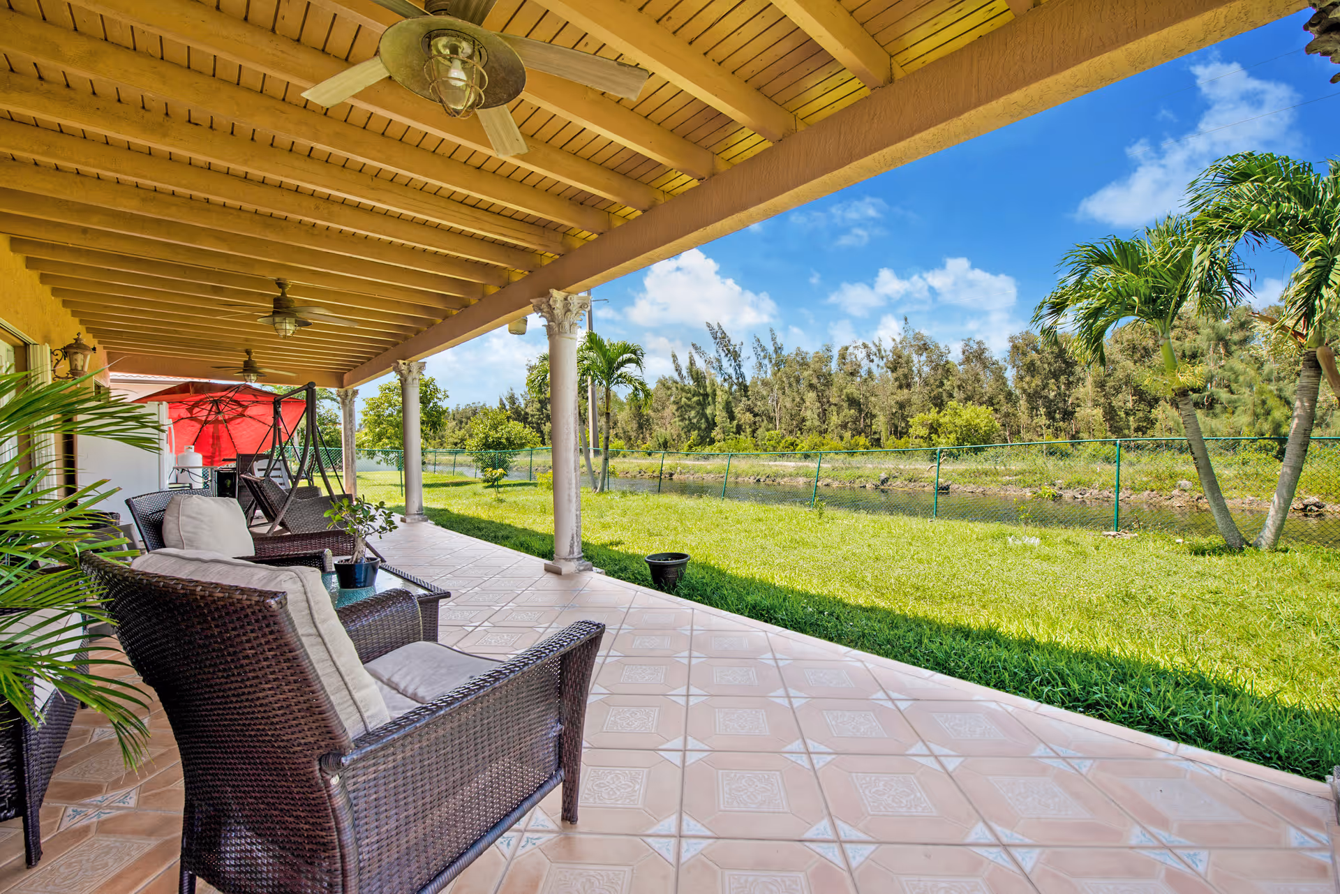 Covered outdoor patio area with wicker chairs and cushions, ceiling fans, and a red umbrella in the background. The patio overlooks a grassy yard with palm trees and a small waterway, under a blue sky with some clouds.