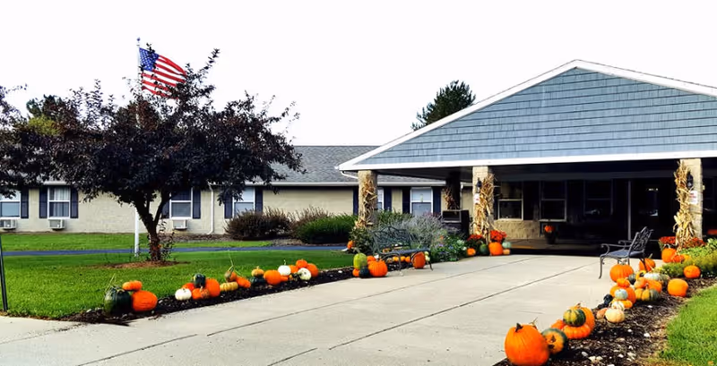 Front entrance of Springcreek Rehabilitation and Nursing Center with a covered driveway and building facade lined with pumpkins and fall decorations and an American flag on the lawn.